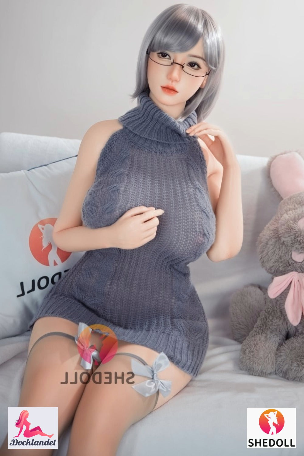 Shedoll sexdocka med kort grå frisyr och glasögon i grå stickad klänning sittandes i soffa bredvid kanin-gosedjur – realistisk silikon-docka från Docklandet