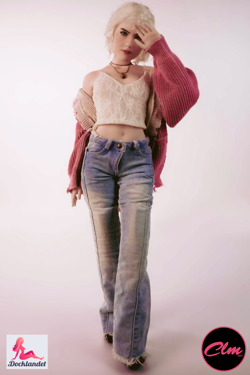 Bambola del sesso Tara realistica mini 60 cm in silicone, bambola bionda in jeans, top corto e giacca rosa, in piedi su sfondo chiaro da studio.